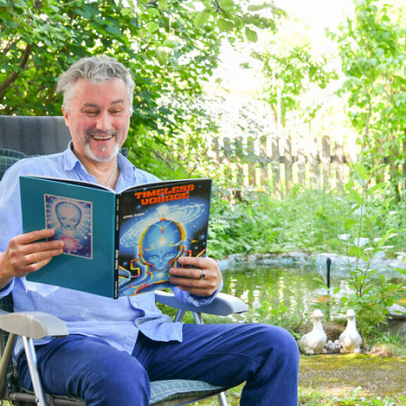 Puutarhassaan istuva Asko tutkii kosmisesta matkustuksesta kertovaa kirjaa suihkulähteen äärellä. Puutarhassaan istuva Asko tutkii kosmisesta matkustuksesta kertovaa kirjaa suihkulähteen äärellä.